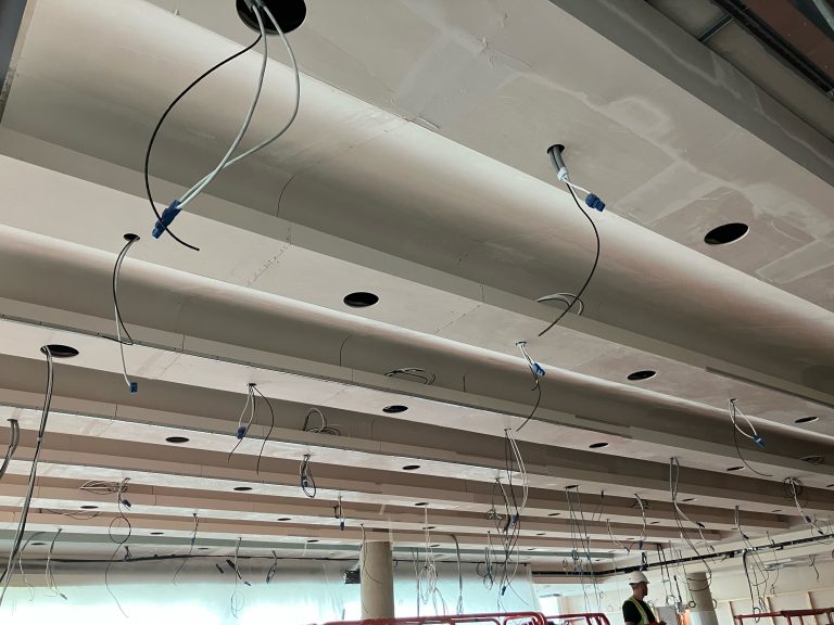 Acoustic wave cassette ceiling installation in progress at Cambridge auditorium, showing sequenced panel installation around structural columns