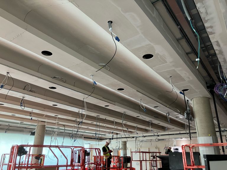 Cambridge auditorium acoustic ceiling cassettes spanning full width of space, with columns and installation team visible